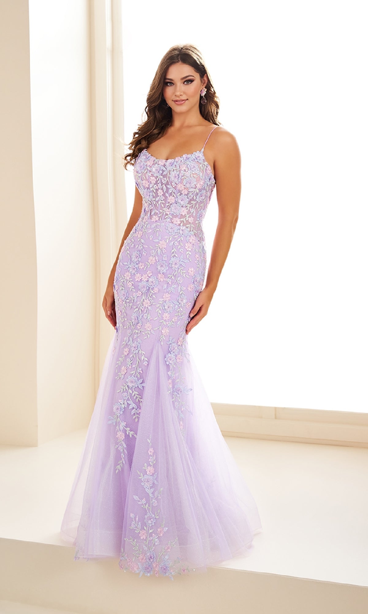 A woman stands indoors wearing the Ellie Wilde EW36090, a fitted lavender mermaid prom dress with floral lace appliques and thin straps.